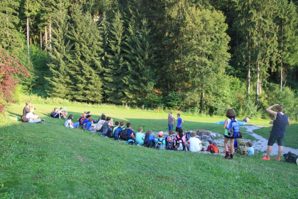 Rund zwei Dutzend Kinder und Jugendliche Sitzen am Waldrand im Kreis und werden von einem Detektiven fürs Schatzsuchen instruiert.